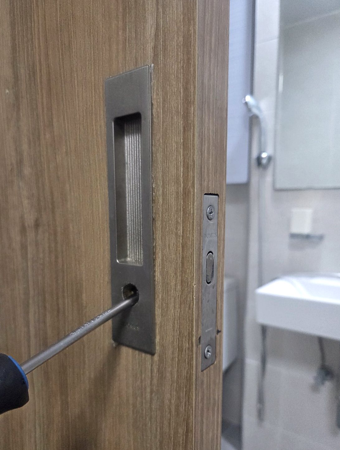 동안구 신촌동 출장 — 나무결 문에 박은 직사각 매립 손잡이와 아래 나사 구멍에 꽂은 드라이버, 문 옆면의 걸쇠 판, 뒤로 보이는 욕실 세면대